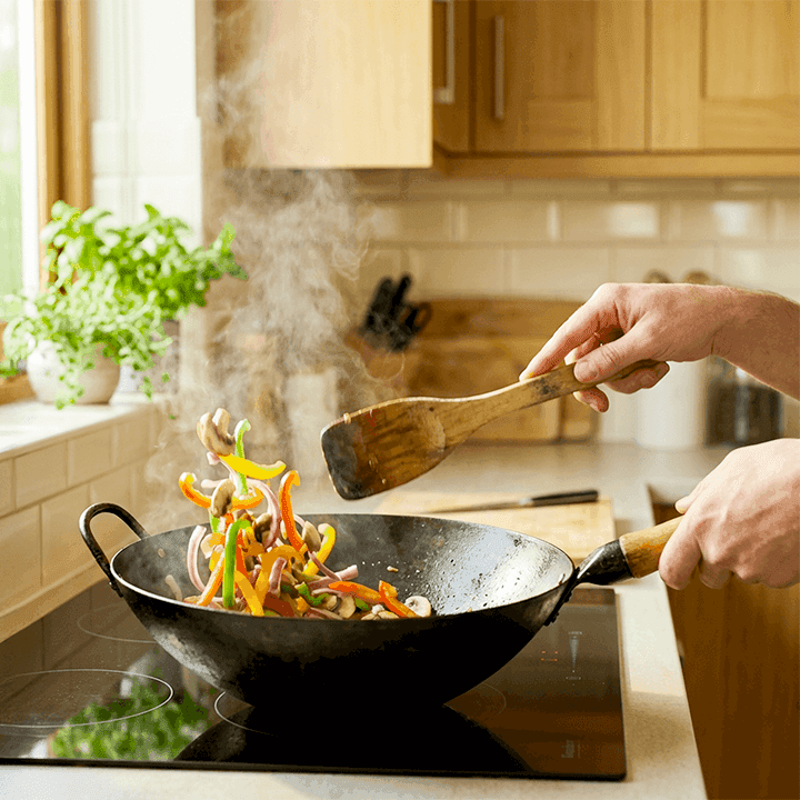 Person frying fajitas consisting of colourful sliced peppers, onions, and mushrooms in a hot wok on a modern kitchen hob, with steam rising and wooden utensils in use.