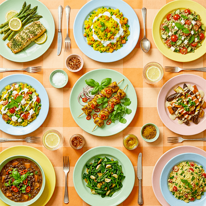 Overhead view of a colourful spread of plated meals on a checkered tablecloth, including grilled salmon with asparagus, spiced rice dishes, fresh salads, vegetable sides, and skewered proteins, arranged with cutlery and small bowls of seasoning.